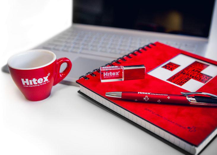 Branded merchandise using Hitex Logo. Espresso cup, pen and notebook in red. Laptop on the background.